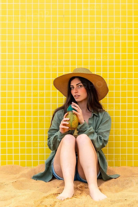 Young woman sitting on the sand, wearing a straw hat and casual clothes, holding a pineapple shaped drink, relaxing against a vibrant yellow tiled wall and embracing summer vacation