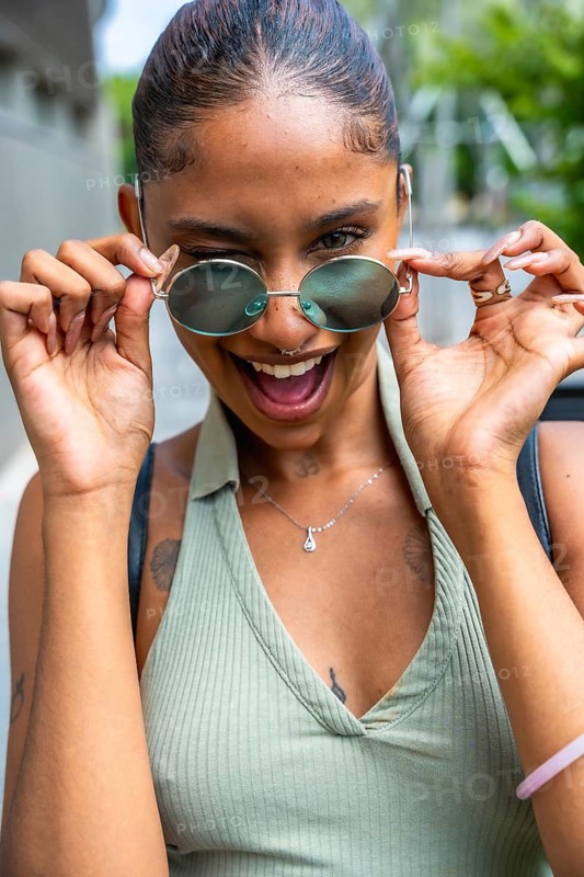 Young woman with tattoos and piercings joyfully gazing over her round sunglasses, expressing happiness and a playful attitude in an urban setting