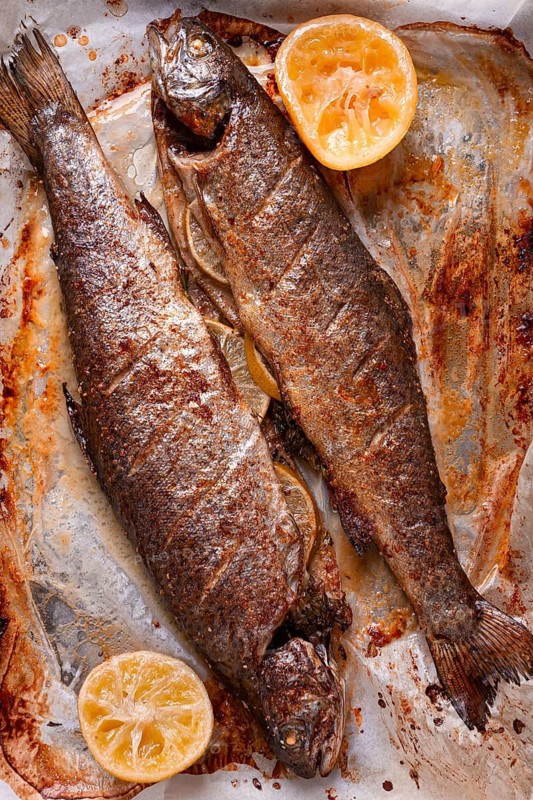 Two fish are being cooked on a baking sheet in a home kitchen. Lemon slices are placed beside the fish to add flavor. The dish shows grill marks and a brown color from baking