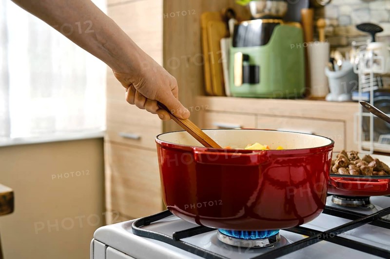 A person stirs a pot on a stove in a kitchen. The pot contains vegetables and is on a gas burner. Light comes in through a window, adding warmth to the cooking process