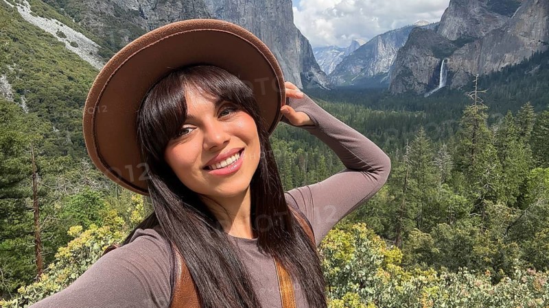 Woman smiling and wearing a hat, capturing a selfie moment in yosemite valley with lower yosemite fall and granite mountains in the background, experiencing the joy of travel and nature