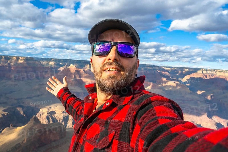 Bearded man wearing sunglasses, a cap, and a plaid shirt, smiling and gesturing towards the vast landscape of the grand canyon national park while taking a selfie during his journey