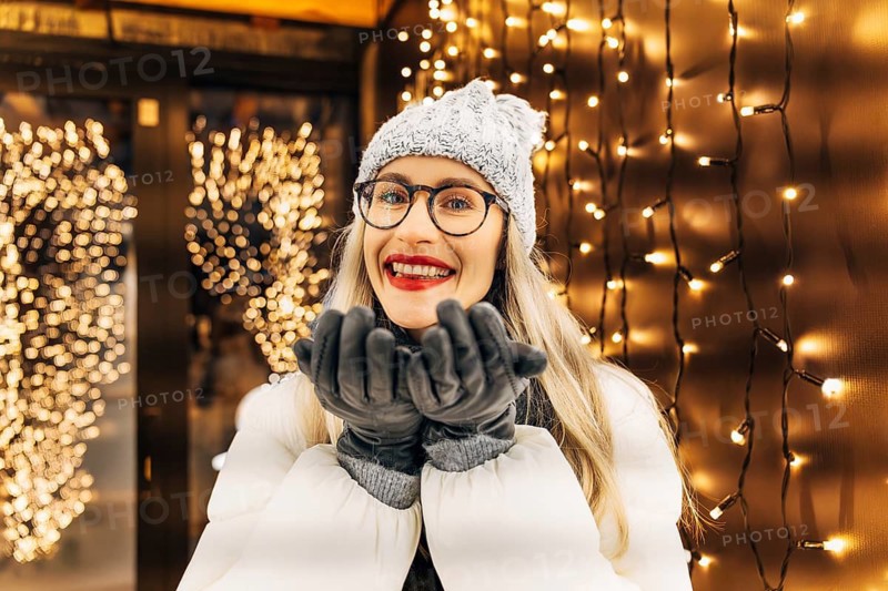 Young woman in winter clothing in front of festive lights. A woman wearing a gray knit hat, glasses, dark scarf, gray gloves and a white winter coat stands outdoors at night with her hands held near her face. Warm yellow holiday lights fill the background, creating dense bokeh and a glowing festive atmosphere. The combination of winter clothing, soft illumination and the cozy gesture conveys a strong Christmas and New Year seasonal mood