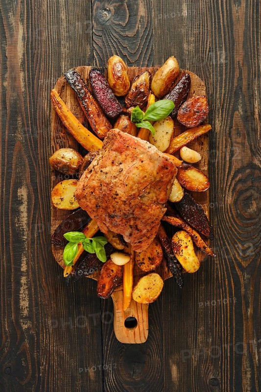 Roasted pork meat on wooden board surrounded by roasted potatoes, carrots and beets. Board is on dark wooden table with natural colors and textures. Meal is ready to serve and enjoy
