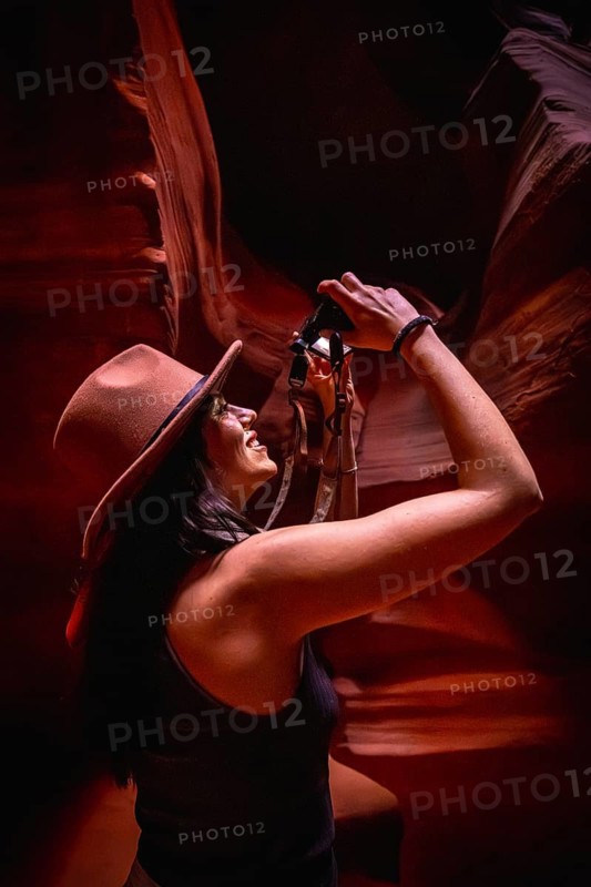 Woman wearing a hat and black tank top, smiling and looking up, holding a camera to photograph the towering, curved red sandstone walls of antelope canyon