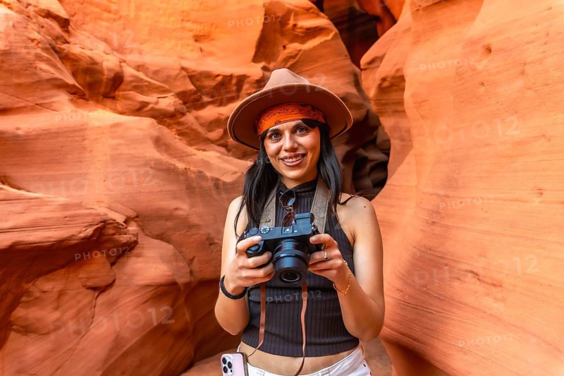 Woman traveler in hat and bandana smiling and holding a camera while photographing the sculpted red sandstone walls and light shafts inside antelope canyon, arizona