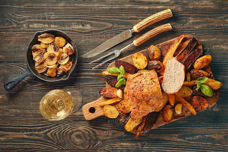 Dinner scene shows roasted pork fillet served on wooden board with various vegetables. Side dish features potatoes. Glass of white wine sits next to food. Focus is on evening meal setting
