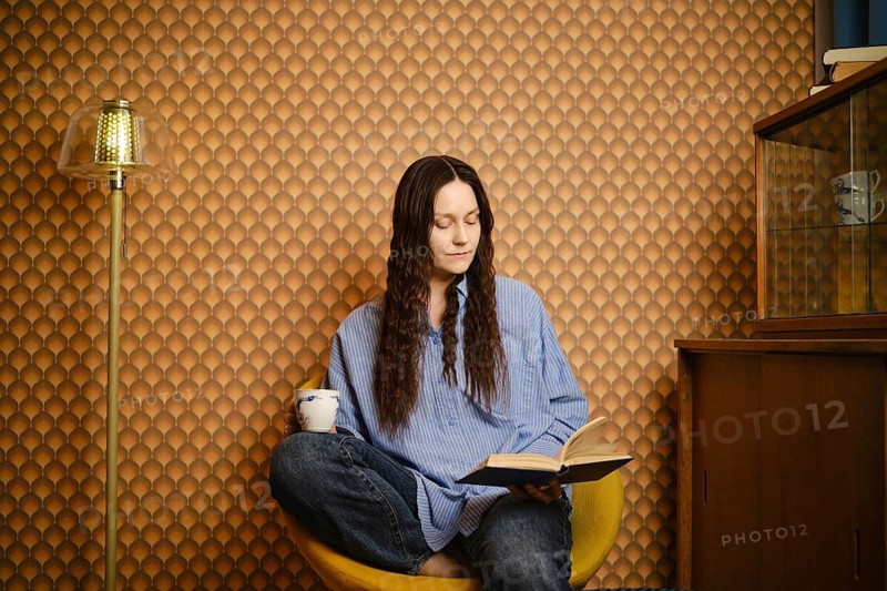 Woman sits in yellow chair, reading book while holding cup. Room has brown patterned wallpaper and display cabinet. Natural light shines in, creating warm atmosphere in afternoon