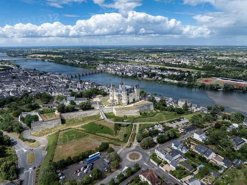 Aerial view of the historic Château de Saumur with its distinctive round corner towers and bright facades, surrounded by well-kept green spaces under a slightly cloudy summer sky, with the town of Saumur on the Loire River, Maine-et-Loire, Pays de la Loire, France