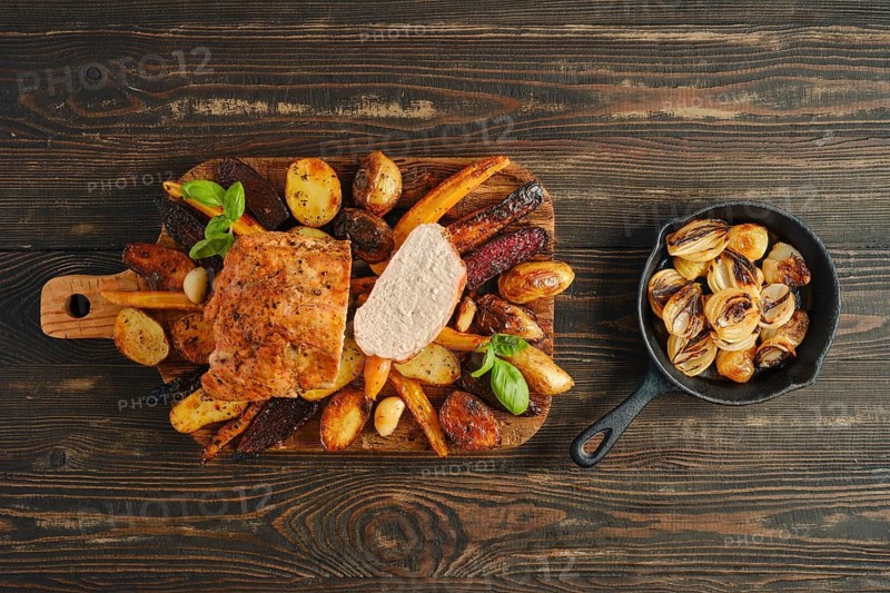 Roasted pork loin on wooden board surrounded by various vegetables. A small pan holds cooked onions. Meal is set on dark wooden table showing rustic kitchen atmosphere