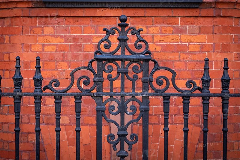 Black, ornate wrought iron fence in front of a brown-red brick wall, Chelsea, London, England, Great Britain