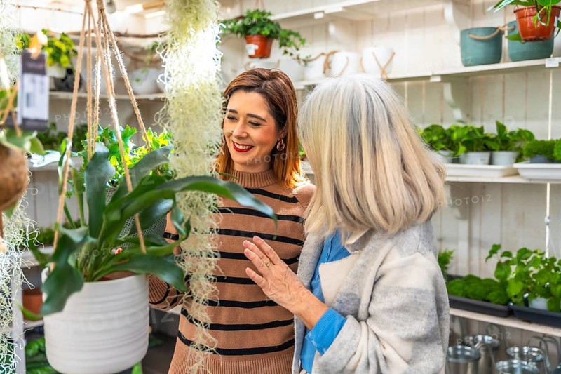 Two multiracial women, one younger and one older, are smiling and engaging while looking at hanging indoor plants in a vibrant garden center, sharing a moment of joy during their shopping experience