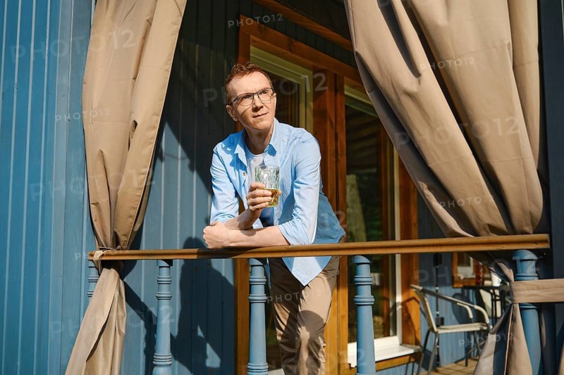 Middle-aged man in blue shirt and beige trousers stands on terrace of wooden cabin looking thoughtful in distance while holding glass in his hand