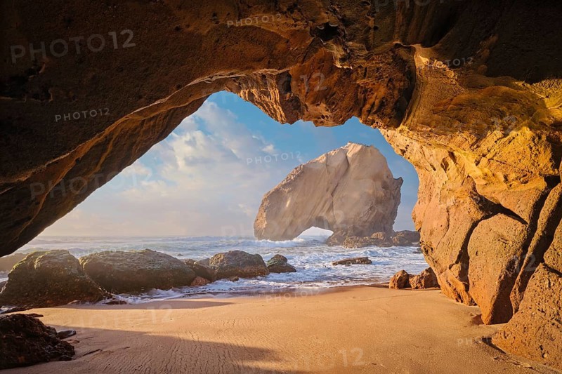 Sunset casts warm colors over Penedo do Guincho rock arch with waves crashing on sandy beach, captured from a grotto cave