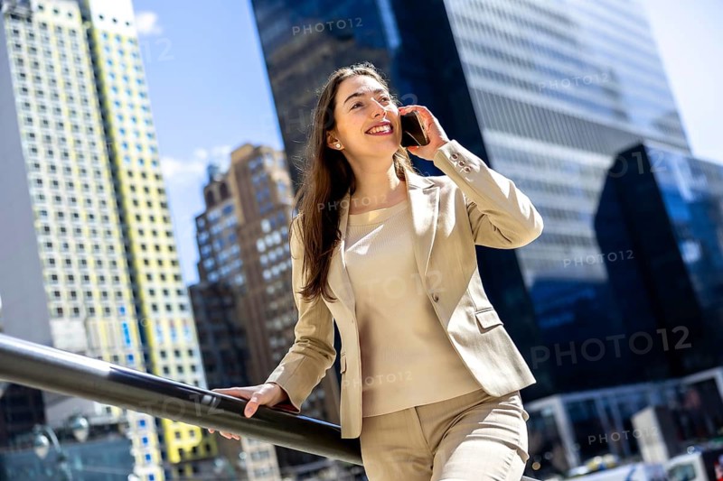 Confident businesswoman talking on her smartphone, smiling while standing outdoors in manhattan with modern skyscrapers creating a vibrant blue city background