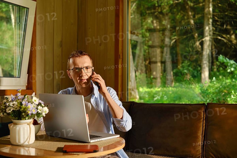 Middle-aged man sits at table in wooden cabin, talking on phone while using laptop. He solves problems over the phone remotely. Green trees visible through windows. Sunlight shines inside creating bright atmosphere during daytime