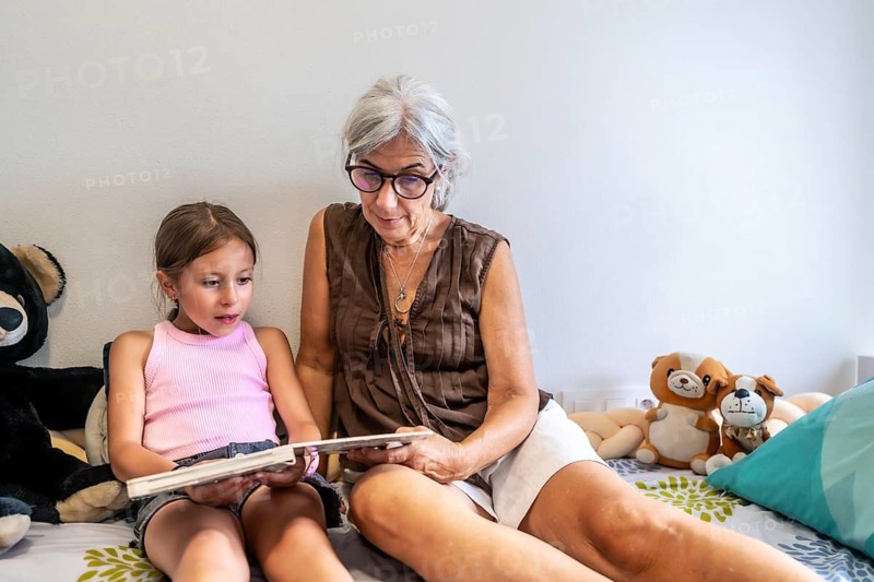 Grandmother is sitting next to her granddaughter, both enjoying a storybook while relaxing on the bed, sharing a moment of intergenerational connection and learning