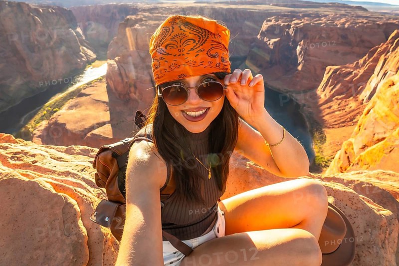 Young woman traveler smiling at the camera while adjusting sunglasses, wearing an orange bandana and sitting on a rock overlooking scenic horseshoe bend in page, arizona on a sunny day