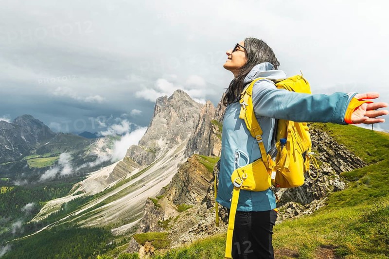 Happy female traveler yellow backpack standing outstretched arms mountain ridge line Seceda South Tyrol Dolomites Italy looking up feeling free peaceful active lifestyle outdoor vacation