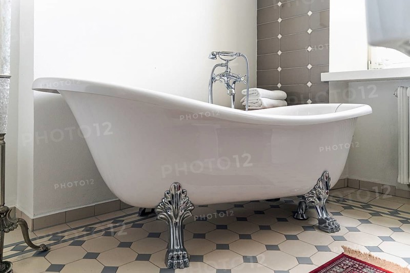A white clawfoot bathtub with ornate silver legs stands in a stylish bathroom. A chrome faucet with a handheld shower is mounted on the tub, and folded white towels rest behind it. The floor features beige and gray hexagonal tiles, while the wall has gray rectangular tiles with small white accents. A window, radiator and part of a red rug complete the classic interior