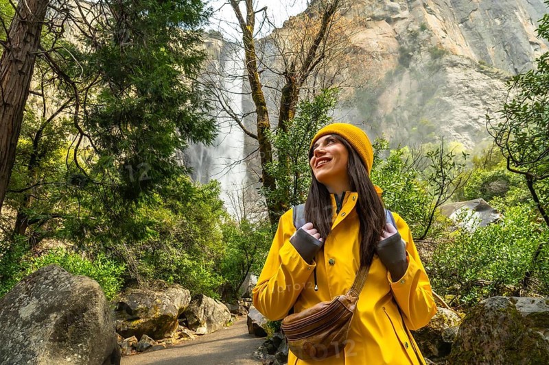 Woman standing on a trail smiling and looking up while hiking through a lush yosemite forest, bridalveil fall and granite cliffs towering in the sunlit background