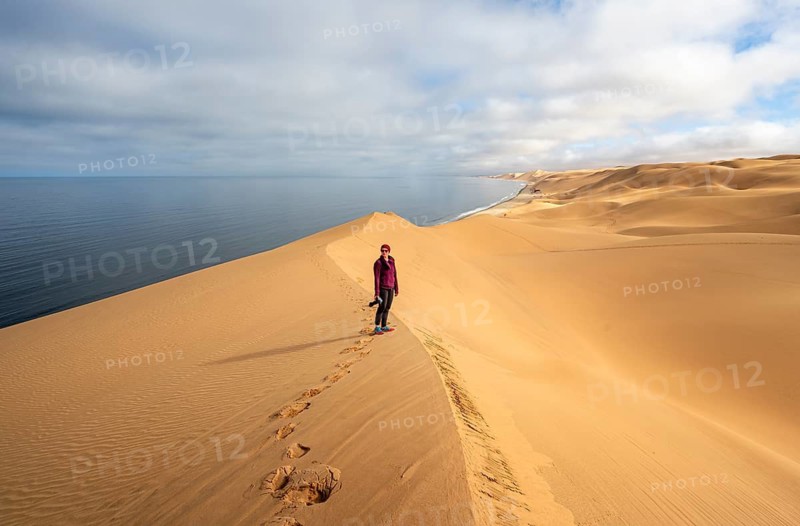 Young woman standing on a sand dune in front of impressive desert landscape and sea, Atlantic coast and Namib desert, Sandwich Harbour, Namib Naukluft Park, Namibia