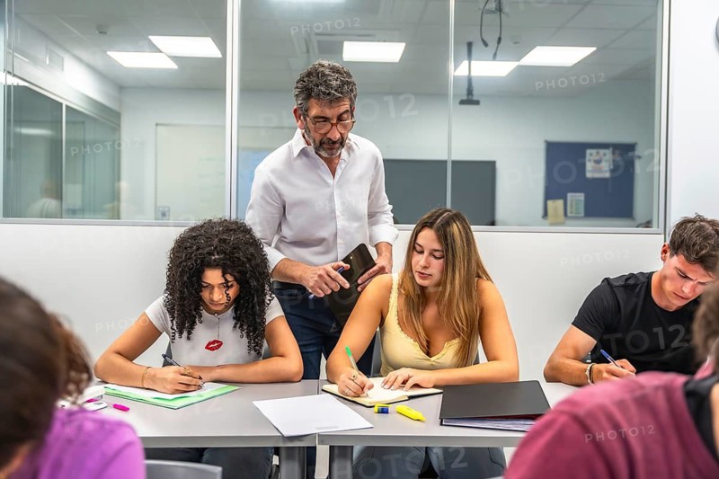 Teacher standing over students, helping with a test while they focus intently on writing answers, demonstrating support and guidance in an academic setting