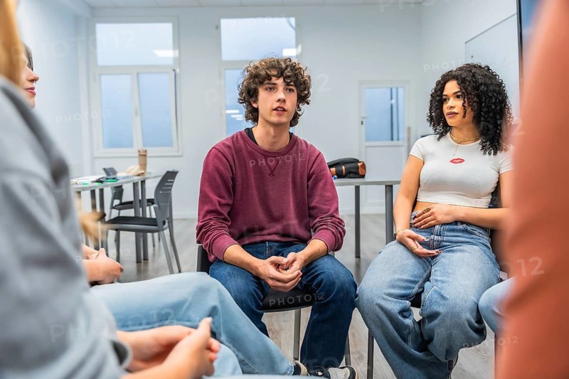 Young adults seated in a circle sharing experiences and listening attentively during a support group session in a bright, modern classroom focused on mental wellness and connection