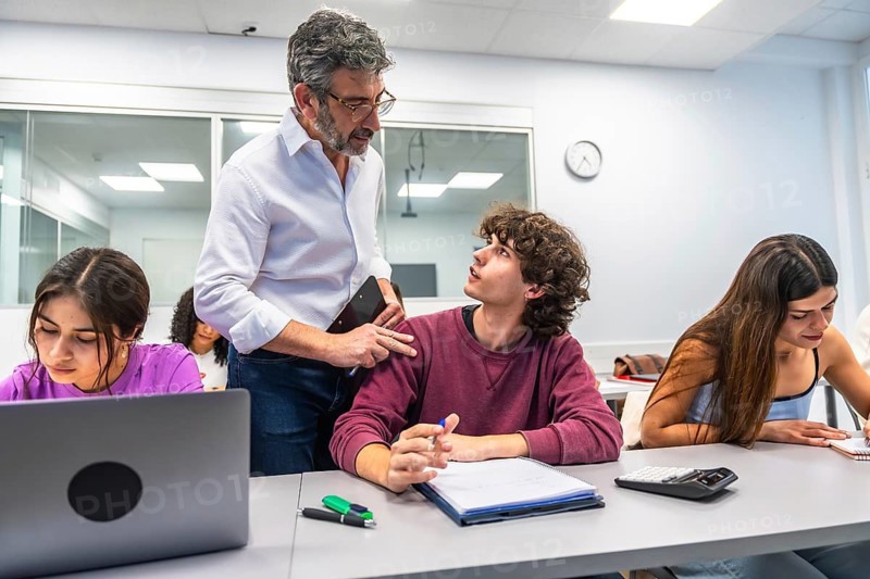 Professor providing individualized assistance and mentorship to a young university student in a diverse college classroom during a lesson, fostering an engaging learning environment