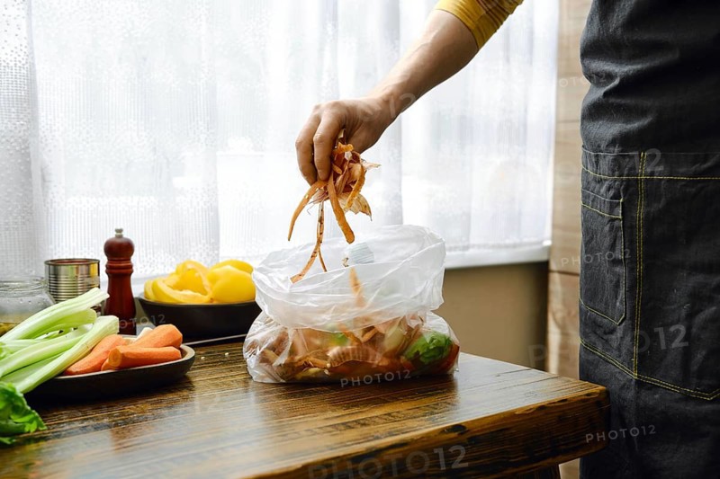 A man is standing in a home kitchen, adding vegetable scraps to a clear bag to add the waste to the compost later. The light from a window shines on the table where fresh vegetables are present