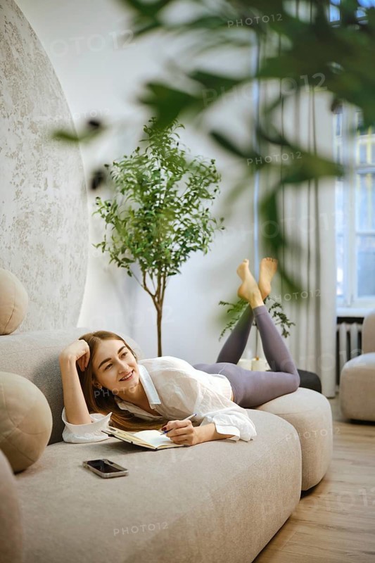 Dreamy woman lies on a soft sofa in her living room, writing in a notebook. She has a relaxed expression and is dressed casually. Bright light enters through a window, creating a warm atmosphere