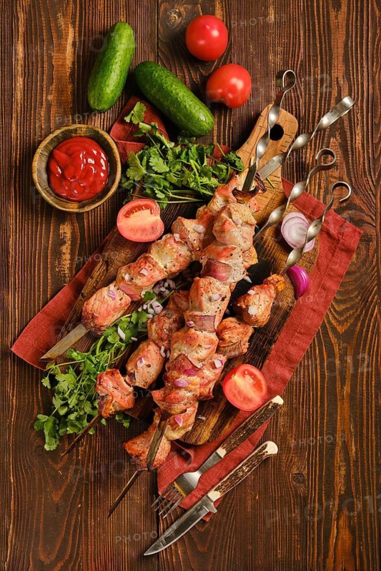 Top view of juicy shish kebab on a wooden board. Fresh tomatoes, cucumbers, and herbs surround the meat. A bowl of sauce is also on the board. This scene is set on a rustic wooden table