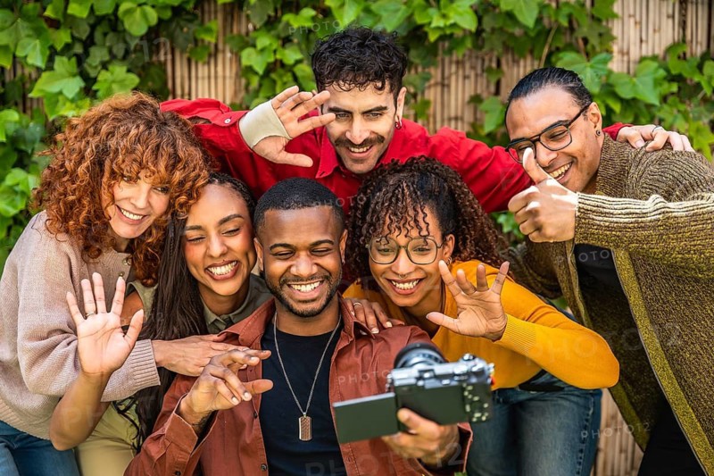 Diverse group of smiling friends creating social media content together with a camera, sharing happiness and connection in a supportive outdoors setting with green foliage