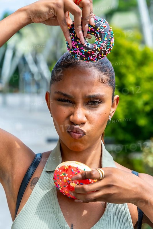 Young woman with a playful expression balancing chocolate and strawberry iced donuts with colorful sprinkles on her head and chest, having fun outdoors