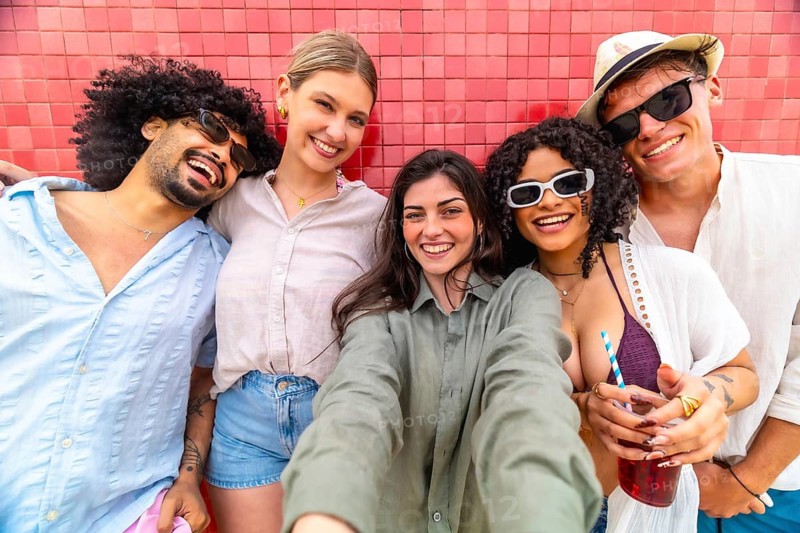 Diverse group of smiling young friends leaning against a red tiled wall, taking a selfie together, celebrating friendship and enjoying their summer vacation