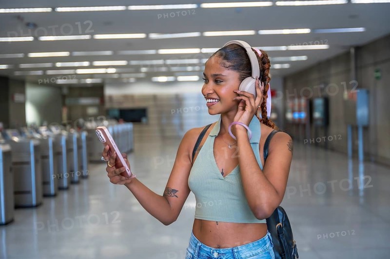 Young woman with headphones and a backpack standing in a subway station, smiling while listening to music and holding her smartphone on her commute to work or travel