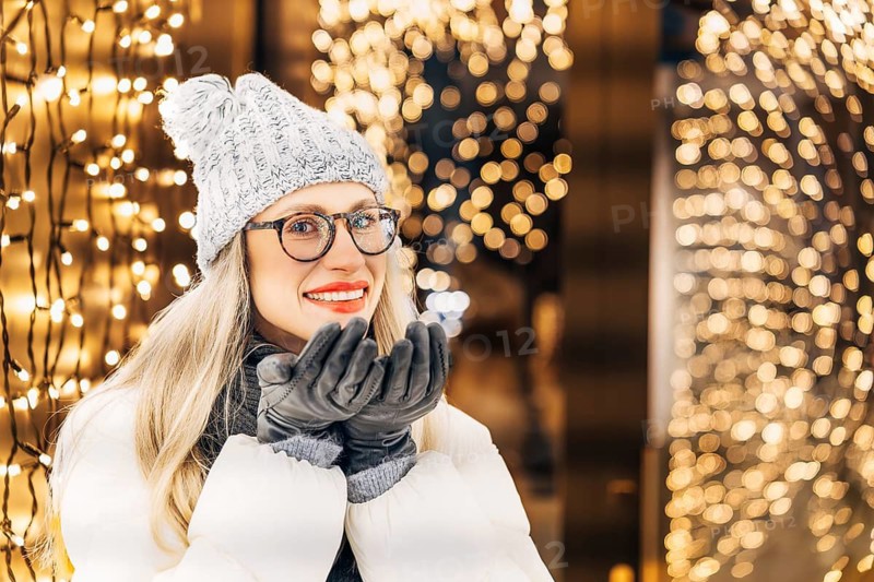 Happy woman in glasses making a wish in winter getting ready to blow on her palms. A woman wearing a gray knit hat, glasses, dark scarf, gray gloves and a white winter coat stands outdoors at night with her hands held near her face. Warm yellow holiday lights fill the background, creating dense bokeh and a glowing festive atmosphere. The combination of winter clothing, soft illumination and the cozy gesture conveys a strong Christmas and New Year seasonal mood
