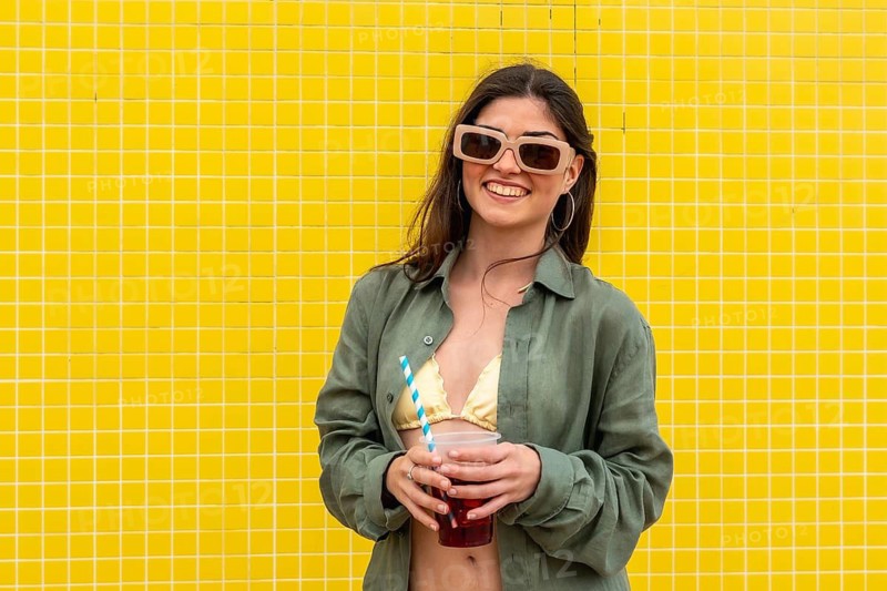 Young woman smiling, wearing sunglasses and a casual shirt over a swimsuit, holding a refreshing drink with a straw against a vibrant yellow tiled wall, embodying summer fashion and leisure