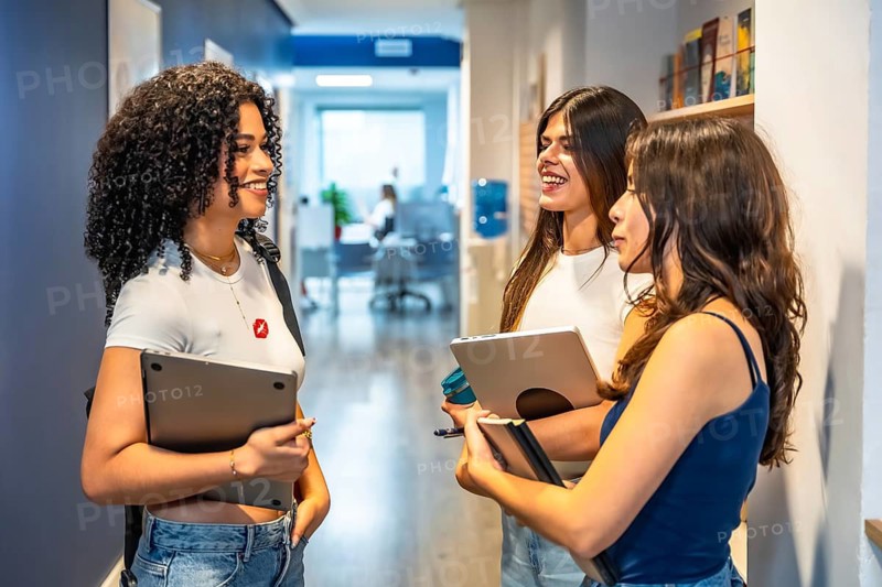 Smiling female university students holding laptops and notebooks, interacting and discussing during a break in a modern school hallway, symbolizing education, teamwork, and campus life