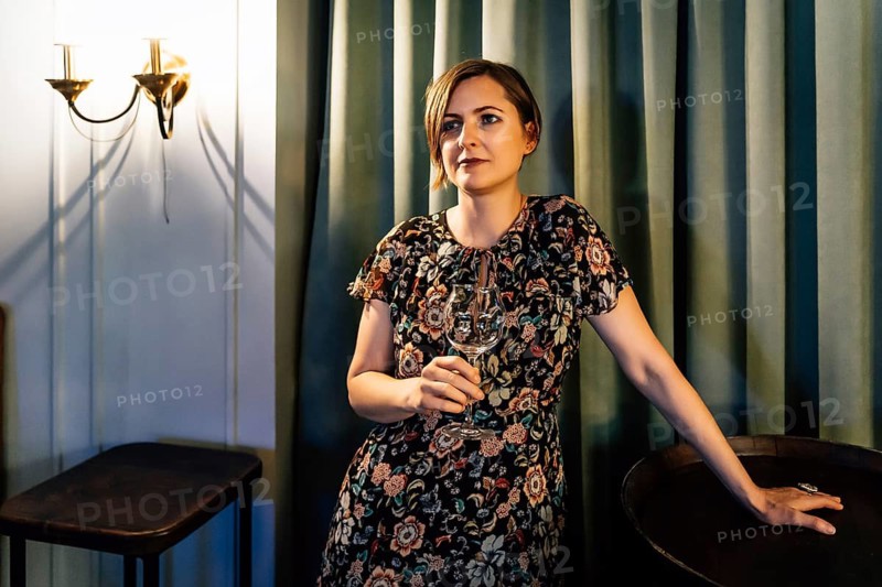 A blonde in a dress with a wine glass stands near a wine barrel. A woman in a dark floral dress stands in a warm bar interior holding a wine glass while resting her hand on a round table. Soft lighting, wall sconces, and tall curtains create a cozy hospitality atmosphere suitable for wine tasting, restaurant promotion, or evening event marketing