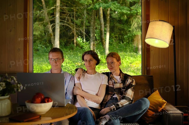 Family sit closely on couch in wooden cabin and watching series on laptop enjoying time together. Concept of advantage of using online streaming platform subscription