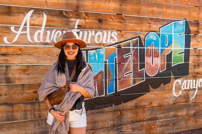 Young woman smiling and wearing a hat, sunglasses, scarf, and white shorts, standing in front of a wooden wall with adventurous antelope canyon text, representing travel and adventure