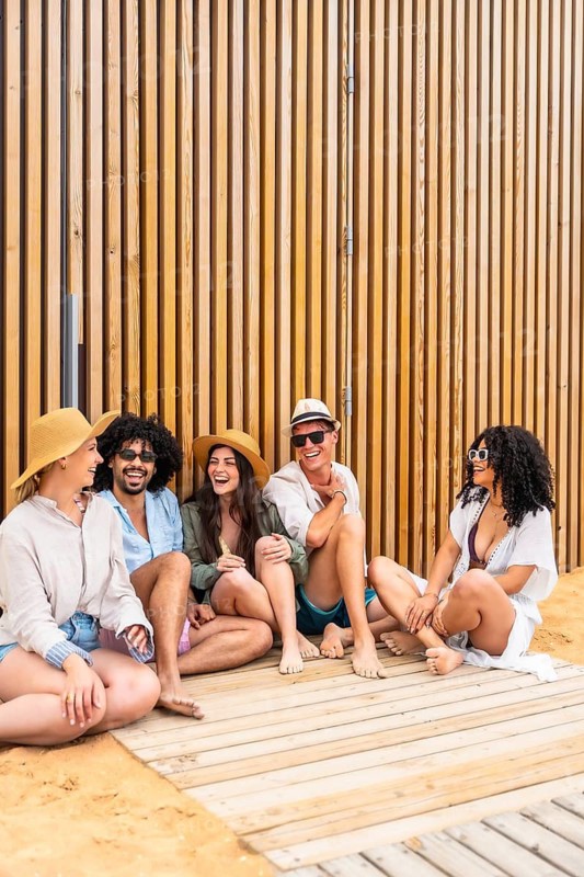 Group of diverse friends in summer clothes laughing and relaxing on a wooden boardwalk at the beach, enjoying sunshine, casual conversation, bare feet and carefree vacation vibes