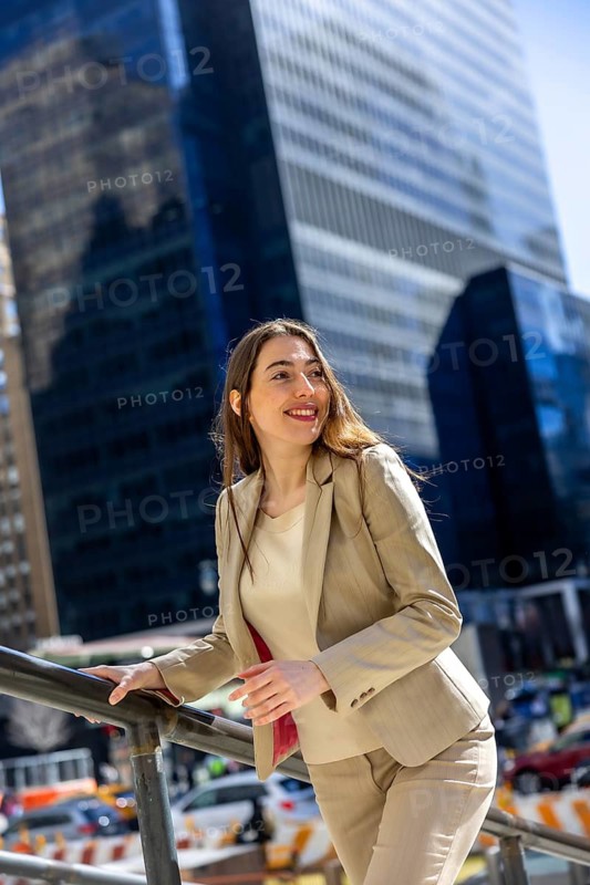 Professional woman in a neat suit is smiling, looking away with a hopeful expression while standing in an urban setting with skyscrapers in the background on a sunny day