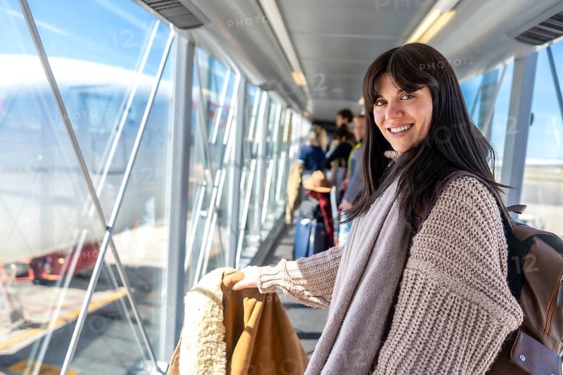 Young woman smiling, ready for travel and vacation, boarding an airplane through a jet bridge with other passengers at the airport terminal, anticipating an upcoming flight