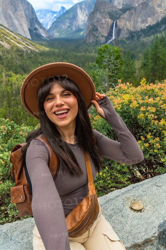 Smiling woman tourist wearing a hat and backpack, happy and excited, experiencing the majestic natural beauty of yosemite valley, with bridalveil falls and el capitan in the background