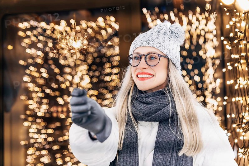 Young happy woman with a sparkler in her hand on the background of glowing garlands. A woman wearing a gray knit hat, glasses, dark scarf, gray gloves and a white winter coat holds a lit sparkler that emits bright sparks. Warm yellow holiday lights fill the background, creating a glowing festive atmosphere. The combination of winter clothing, illuminated decorations and the active sparkler adds a strong celebratory Christmas and New Year mood