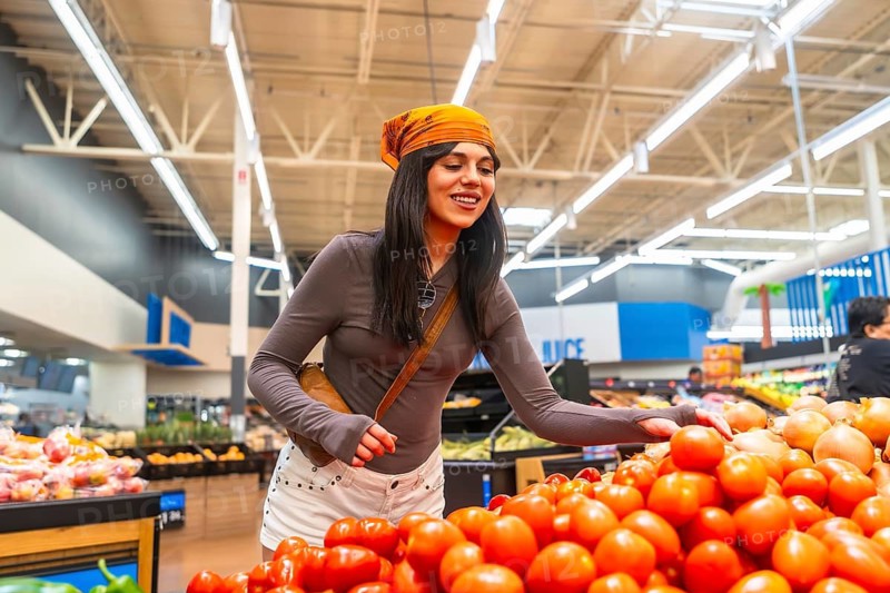 Young woman wearing a headscarf and brown long sleeve shirt selecting ripe red tomatoes from a large display in the produce section of a brightly lit grocery store