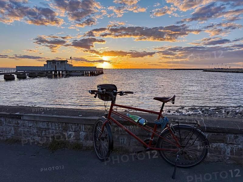 A bicycle is parked in front of a stone wall with a view of the sea at sunset. A pier stretches into water under a colorful sky, Borgholm, Öland, Sweden