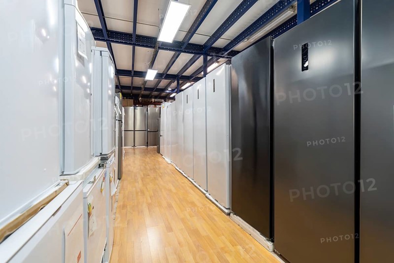 Multiple rows of various refrigerators, including white, stainless steel, and black models, displayed along a wooden floor in a spacious warehouse style home improvement store with overhead lighting